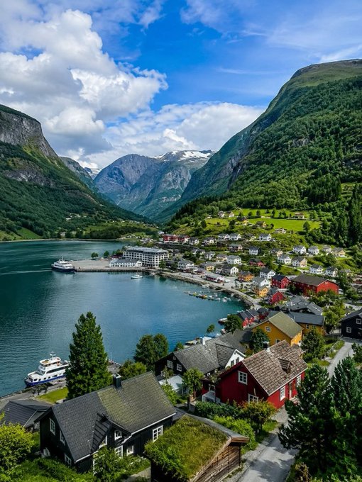 A scenic fjord village—likely Aurlandsfjord in Norway—nestled between lush green mountains and calm blue waters under a partly cloudy sky, with colorful houses and boats dotting the shoreline. The serene, picturesque landscape parallels the post's message of a calm, prosperous Sunday, while the name "$RIVER" reflects the tranquil waterway winding through the valley.