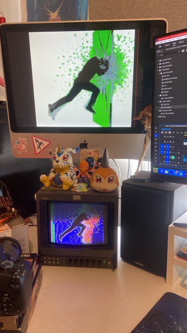 A desk setup featuring an Apple monitor displaying a green and black silhouette graphic, with small figurines and a Sony CRT television below also showing a similar silhouette image in pink and blue.