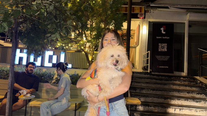A woman holds a fluffy white dog in front of an outdoor venue with a sign reading 'team turkiye', while two people sit on a bench nearby.