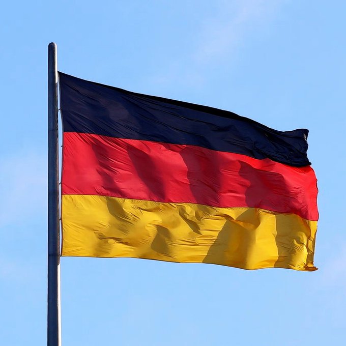 The German national flag (black, red, and gold horizontal stripes) waving on a flagpole against a blue sky.