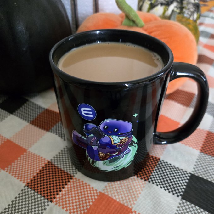 A black coffee mug featuring a cartoon purple creature riding a ghost sits on a plaid tablecloth next to a pumpkin.