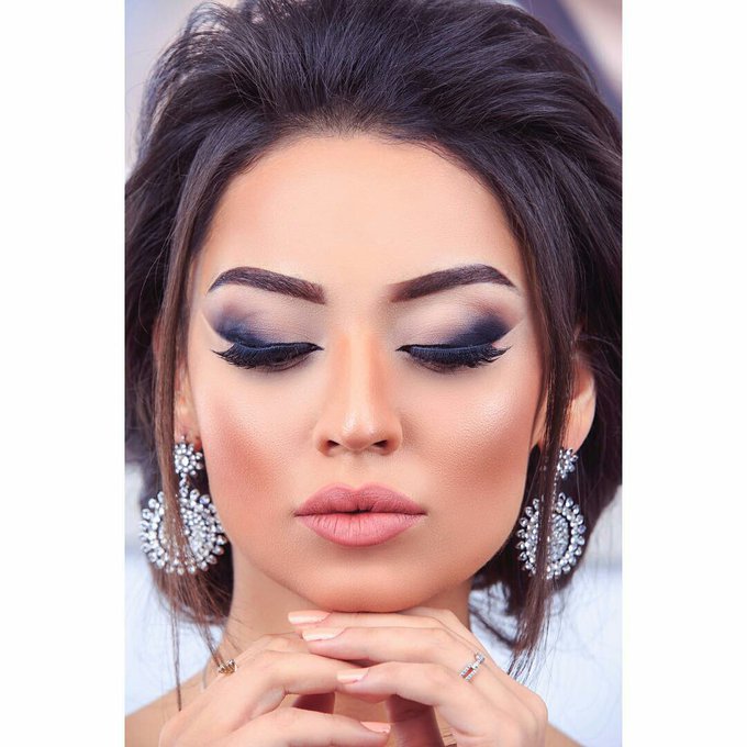 A close-up beauty portrait of a woman with dramatic smoky eye makeup, defined brows, and large ornate silver earrings, with her hands gently resting under her chin.