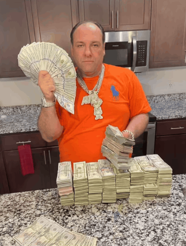 A photograph of a man in an orange shirt and a large silver chain standing in a kitchen, holding and surrounded by stacks of cash.