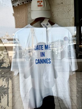 A storefront window display in Cannes featuring a white t-shirt with the text "DATE ME IN CANNES" layered over a sailor hat, a striped collar, and printed photos of a harbor scene.