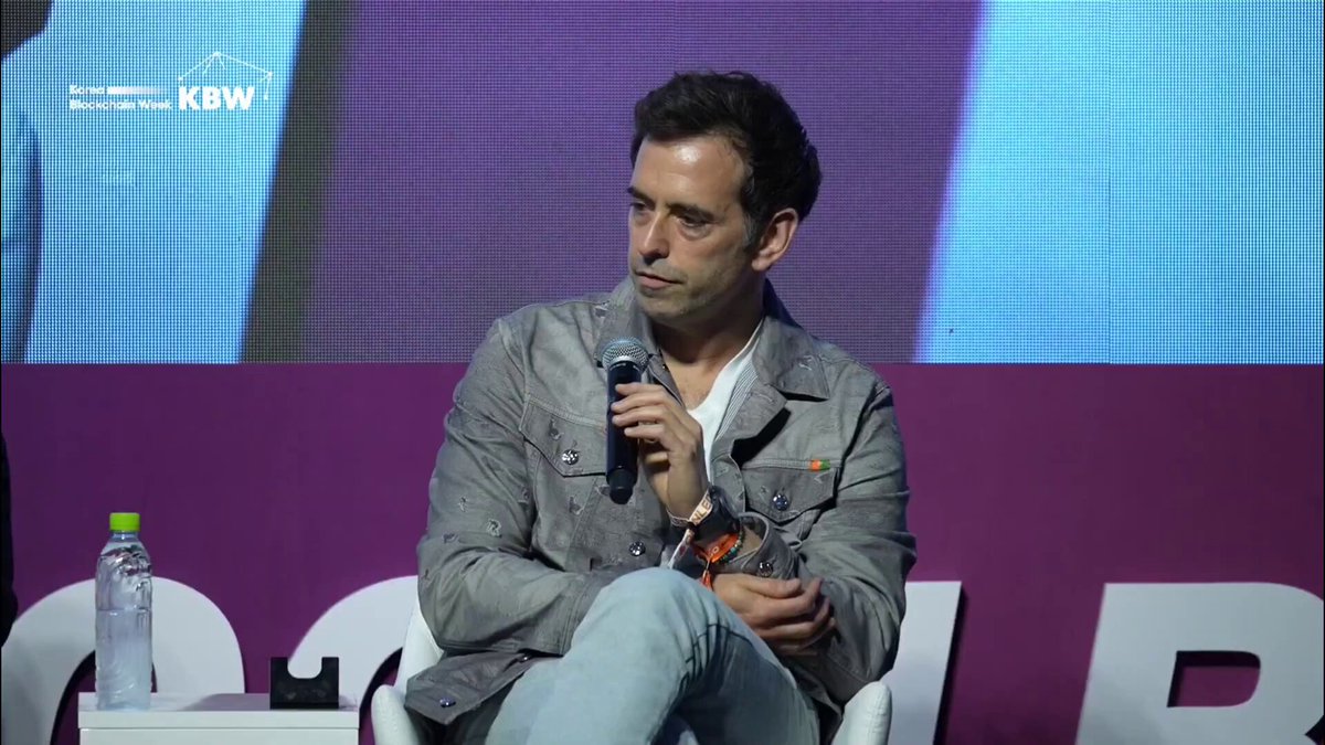 The image shows a man sitting down, holding a microphone, and speaking into it. The background features a purple wall with white text that reads "L1s assumed a homogeneous world. Different countries, different laws, different institutional requirements have shown that to be difficult. Privacy and control as first principles is what'S enabled Canton to work within that reality." This suggests the man might be discussing the challenges of implementing privacy controls in a diverse global context or addressing the complexities of international law and regulations.