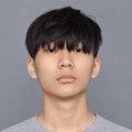 A headshot portrait of a young man with dark hair wearing a gray shirt, photographed against a plain gray background.