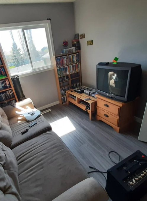 A cozy living room features a brown sectional sofa facing a tube TV on a wooden stand, with a bookshelf filled with DVDs/games beside a bright window, and a black amplifier with cables on the floor in the foreground.