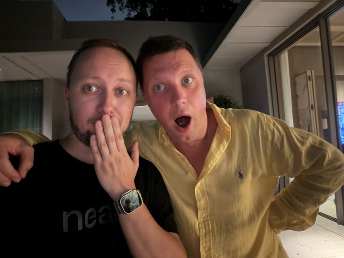Two men pose closely together at night, one with his hand over his mouth wearing a black 'Neam' shirt and Apple Watch, and the other in a yellow button-up shirt making an exaggerated surprised expression.