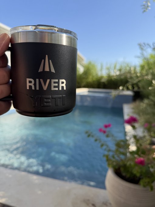 A hand holding a black insulated mug branded "RIVER YETI" in front of a swimming pool and sunny outdoor setting.