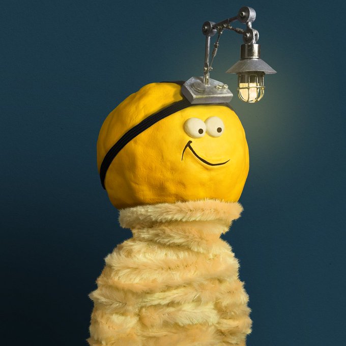 A whimsical sculpture of a yellow smiley-face ball with a miner's lamp attached on top, resting on a fuzzy, mummy-like textured body against a dark blue background.