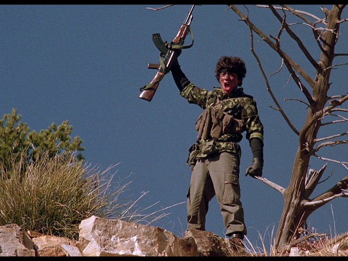 A camouflaged figure in a fur hat triumphantly raises an AK-47 against a clear blue sky while standing atop a rocky outcrop, with a bare tree beside them. The image satirizes the "Colorado is run by communists now" post by humorously framing the gunman as a stereotypical revolutionary militant, evoking Cold War-era anti-communist fears.