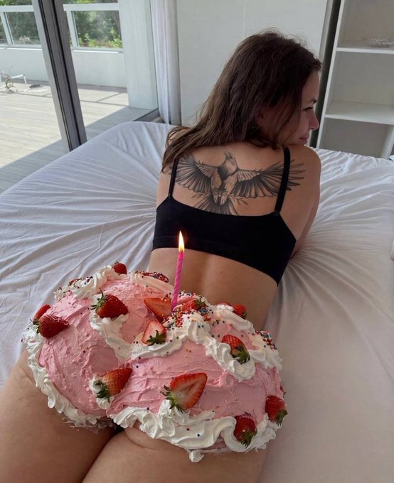 A woman with a large back tattoo of a bird poses on a bed, her body partially covered by a pink cake shaped like her lower half, complete with strawberries and a lit candle.