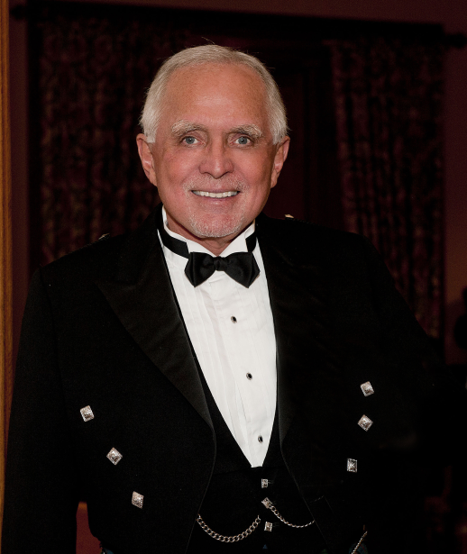 A distinguished older gentleman with white hair and a goatee smiles at the camera while wearing a formal black tuxedo with bow tie, photographed against a dark curtain backdrop. The image appears unrelated to the accompanying post about cryptocurrency investments like AI, CASHCAT, MARSCOIN, and ZCAT, as no text, tickers, or identifiable figures related to these topics are visible.