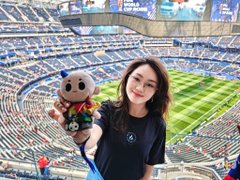 A woman smiles while holding up a small plush mascot toy inside a large stadium, with the green field and crowded stands visible behind her. The post highlights her appearance at the World Cup as a VIP guest sponsored by Huobi (HTX), showcasing the exchange's high-end hospitality experience.