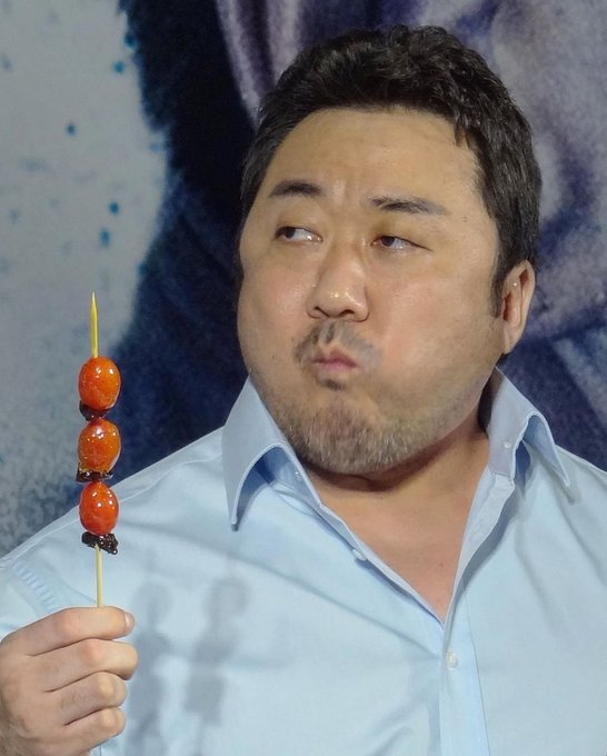 A man in a light blue shirt holds a skewer of candied hawthorn (tanghulu) while making a pouting expression, looking off to the side.