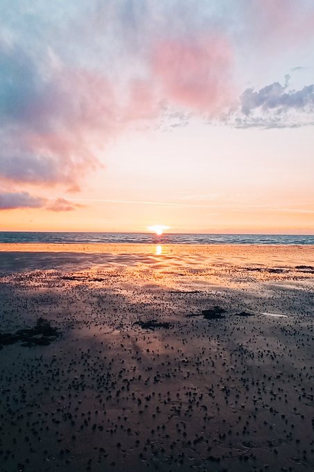 A scenic beach at sunset with pink and orange clouds reflecting on wet sand and tide pools, the sun dipping low on the horizon over calm ocean waters.