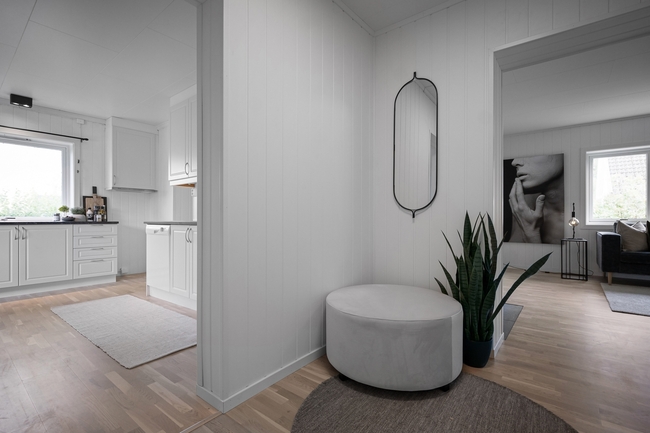 A bright, modern interior with white paneled walls features a round gray ottoman, an oval mirror, and a potted snake plant, with a kitchen visible on the left and a living room with artwork on the right.