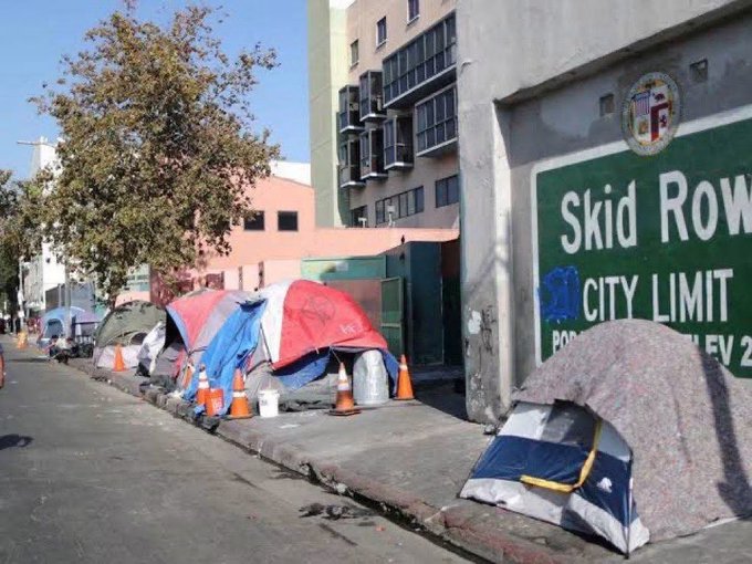 Tents line the sidewalk next to a wall painted with a 'Skid Row City Limit' sign, depicting urban homelessness in Los Angeles.