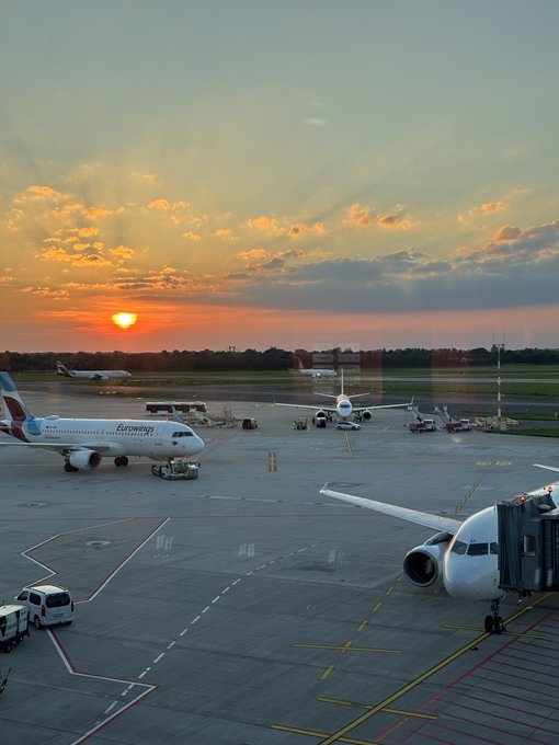 An airport tarmac scene at sunset with several airplanes, including a Eurowings aircraft, parked at gates and taxiing, with a warm, colorful sky in the background.