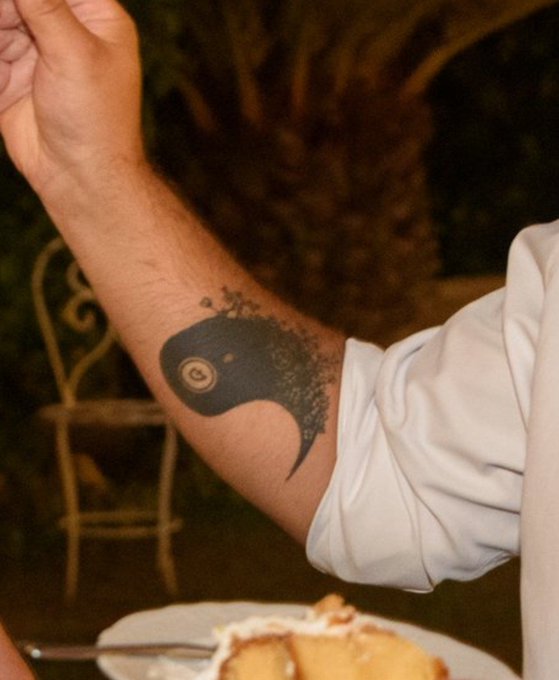 A close-up of a person's forearm displaying a black ink tattoo of a stylized bird or fish-like design with a single visible eye.