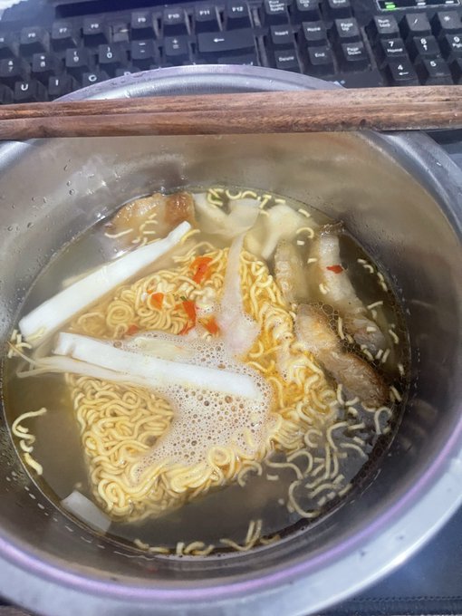 A bowl of instant noodles in broth with chopsticks resting on top, sitting on a desk in front of a black computer keyboard.