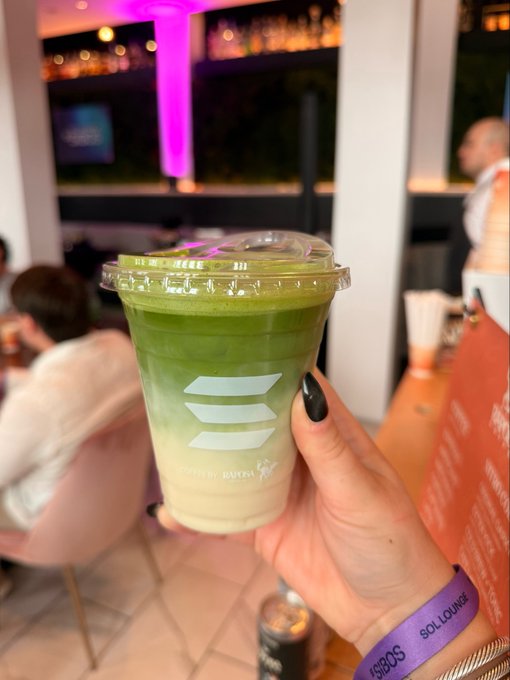 A hand with black nail polish and a purple 'SO LOUNGE' wristband holding a layered green and white matcha-like drink in a clear cup, photographed in a lounge setting.