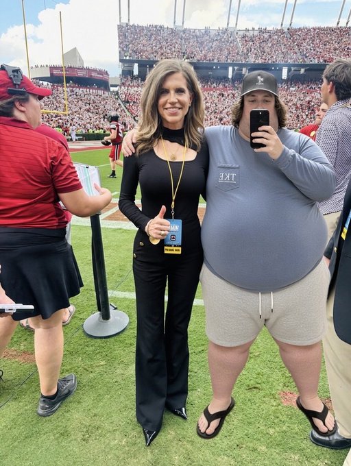 Two people posing at a football stadium - a tall woman in a black outfit with a credential lanyard and a man wearing a black cap, gray t-shirt, and shorts taking a mirror selfie with his phone.