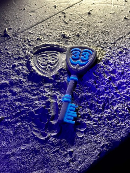 A 3D-rendered key with a blue '33' emblem lying next to a shield crest on a textured, cratered blue surface.