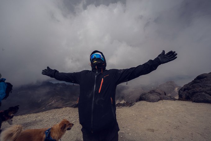 A person in a black jacket, ski mask, and blue goggles stands with arms outstretched on a misty, rocky mountain summit, accompanied by a small brown dog.