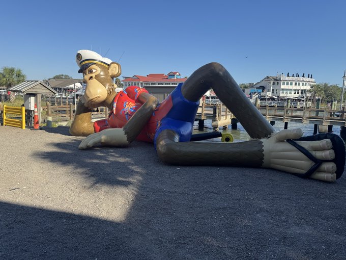 A large monkey statue wearing a captain's hat, red Hawaiian shirt, and blue swim trunks, reclining leisurely on a waterfront dock with a skateboard beside it.