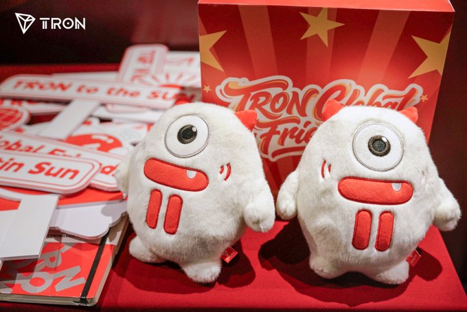 Two white fuzzy plush toys with one eye and red mouths stand in front of a red box with "TRON" branding and star designs, with promotional stickers reading "TRON to the SUN" visible.