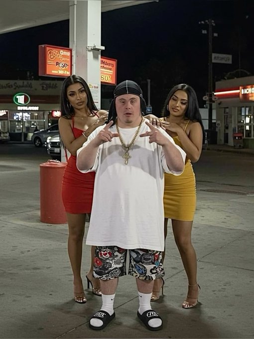A man wearing a durag, oversized white t-shirt, and skull-print shorts poses with two women in form-fitting red and yellow dresses at a gas station at night.