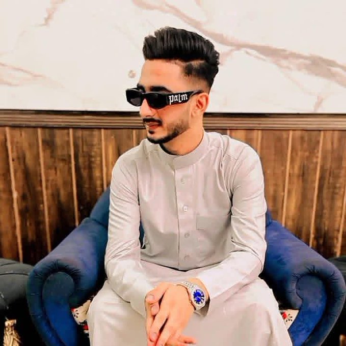 A stylish man with a fade haircut, beard, and rectangular sunglasses featuring the "palm" logo sits on a navy blue tufted couch against a wood-paneled and marble wall, wearing a crisp light gray traditional shalwar kameez and a luxury watch. The confident, put-together pose mirrors the post's upbeat crypto hustle energy—ready to chase "magic internet money" and weekend gains with flair.