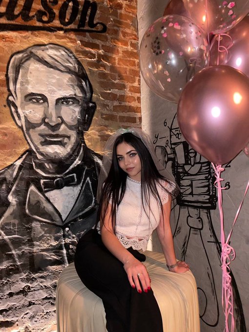 The image features a woman sitting on a stool or bench with her legs crossed, wearing black pants and a white top. She has red nail polish on her fingers and is posing for a picture. The background includes a brick wall with a painting of a man's face, pink balloons, and a sign that reads "Gm frens waiting for a @Tangem ring like it’s the only proposal I need not tryna get wifed up, just trypan hold my bags on my finger stacking sats while looking cute". The woman is positioned in front of this wall with her legs crossed and hands resting on her lap.