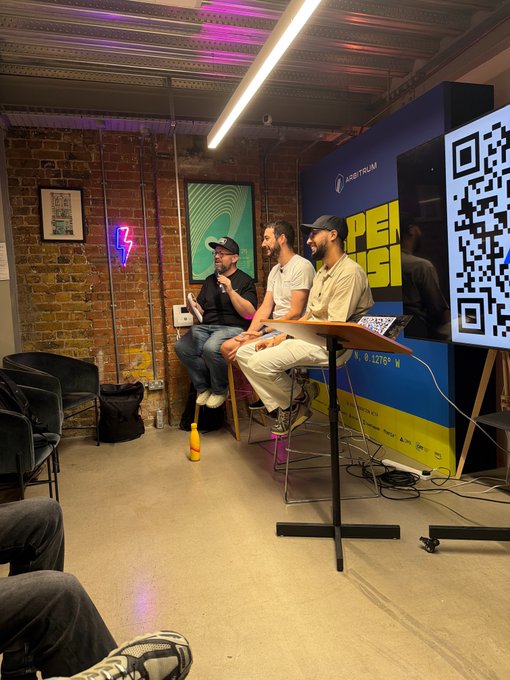 Three men sit on stools at a panel discussion in a brick-walled venue featuring Arbitrum branding and a large QR code, with one speaker holding a microphone while discussing UK crypto builders like xLiquida, who are tokenizing UK government bonds and developing a regulated collateral rail.