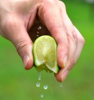 Close-up photograph of hands squeezing a lime half, with juice dripping down against a blurred green background.
