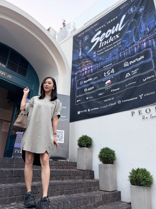 A woman in a gray t-shirt dress poses on the steps in front of a large banner for the "SEOUL INDEX" event (presented by Google Cloud, CoinMarketCap, and Strategy, with Title sponsor t54 and Portals as a major partner), looking like she's waiting for friends to arrive at the venue.