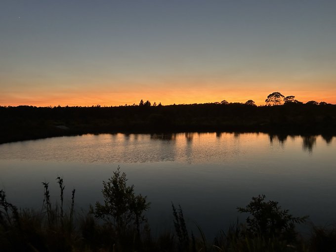 A serene sunset over a calm lake, with silhouettes of trees reflected in the water beneath a gradient orange and blue sky.