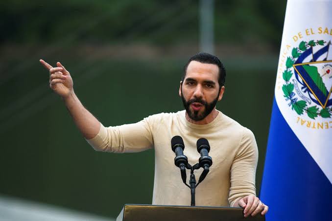 A bearded man in a beige sweater speaks passionately at a podium with two microphones, gesturing with his right hand beside a flag bearing the "REPÚBLICA DE EL SALVADOR" coat of arms, appearing to commemorate the 5-year anniversary of El Salvador's historic 400 BTC purchase as the first nation to adopt Bitcoin.