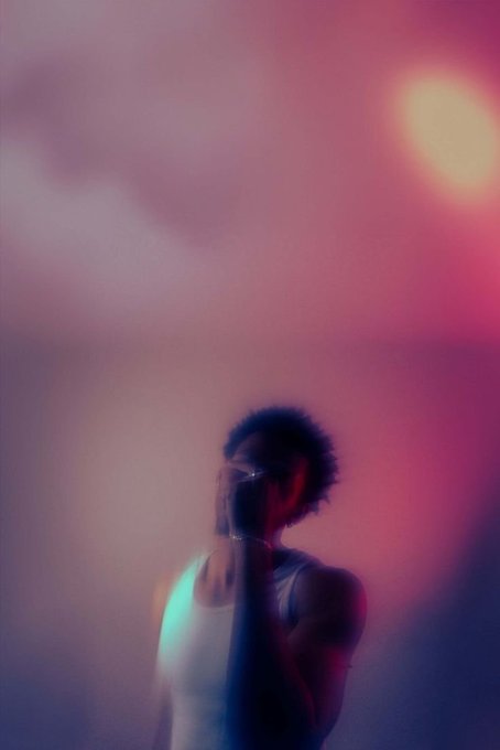 A blurry, atmospheric portrait of a person with short dark hair and glasses wearing a white tank top, bathed in pink, red, and teal lighting.