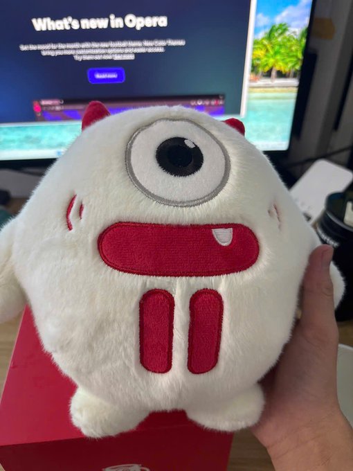 A hand holds a plush toy featuring a single large eye, small red horns, and a red mouth design, sitting on a red box in front of a monitor displaying "What's new in Opera" with a $2500 TRX pool promotion, matching the post's mention of a limited-edition TRON bear giveaway.