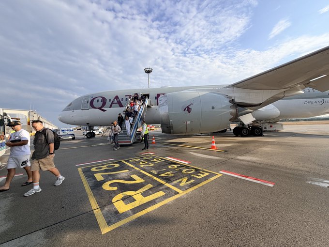 Passengers deplane a Qatar Airways aircraft onto the tarmac, with 'NE B12 STOP R757B' markings painted on the ground.