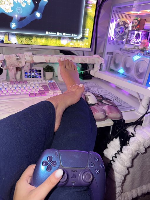 A first-person view shows a person holding a blue PlayStation 5 DualSense controller, with their bare feet resting near a pink keyboard and monitor displaying a game.