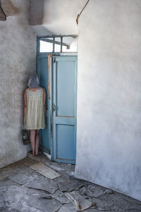 A haunting photograph of a young girl in a pale yellow dress and headscarf standing barefoot before a weathered blue door, surrounded by rough, crumbling walls and debris on the floor.