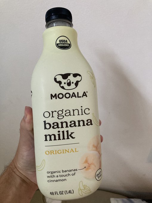 A hand holds a 48 FL OZ (1.4L) bottle of MOOALA Organic Banana Milk, which is USDA Organic and features a 'with a touch of cinnamon' label.