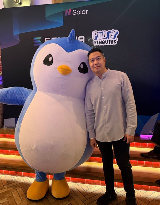 A man in a light blue button-up shirt stands next to a large Pudgy Penguins mascot in front of a branded "Solar Huddle" and "Pudgy Penguins" backdrop at an event in Singapore. The image aligns with the post's caption about attending the Solana Huddle in Singapore alongside the Pudgy Penguins team.
