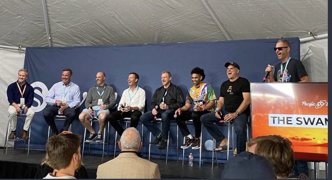 A panel of seven speakers sits on stage at a Pacific Bitcoin event, with a moderator holding a microphone addressing the audience from behind a podium displaying "THE SWAN" branding. The lively gathering, with panelists smiling and laughing, reflects the celebratory spirit of the Pacific Bitcoin conference.