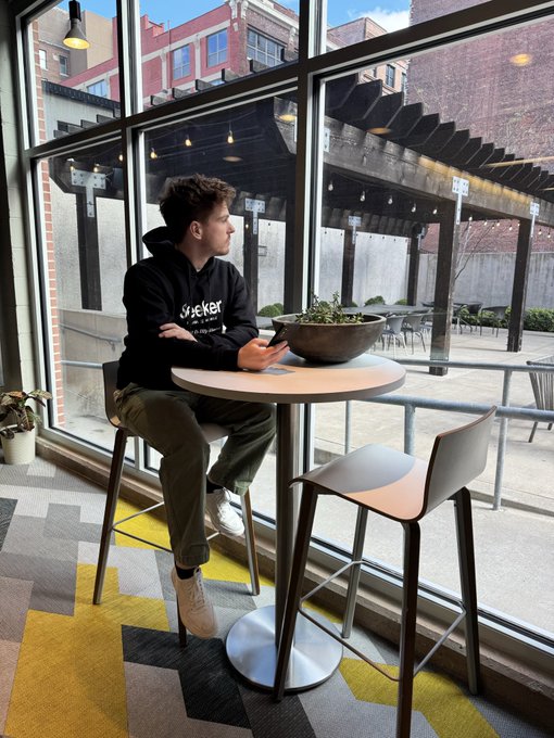 A young man wearing a black 'Live Nicker' hoodie sitting alone at a high table in a cafe, looking at his phone while gazing out a large window at a brick building.