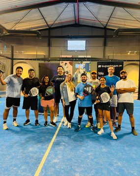 The image features a group of people posing for a photo with their tennis rackets, standing together on a blue court. There are at least nine individuals in the picture, all holding tennis rackets and smiling for the camera. The background includes a large window that allows natural light into the room, as well as some posters hanging on the wall. One of these posters reads "padel done the solana way." This suggests that they might be participating in an event or gathering related to tennis or outdoor sports.