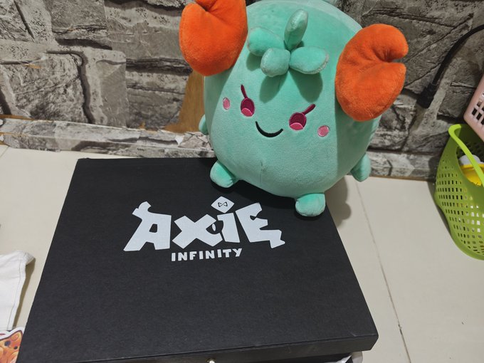 A teal plush toy of the Axie character 'Aqua' from the game Axie Infinity, sitting on a black 'AXIE INFINITY' branded box against a stone wall.
